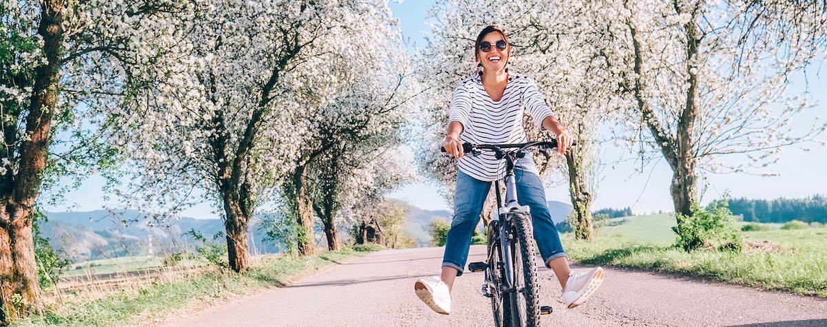 A woman in a striped shirt riding a bike on a dirt road lined with trees and grass during a beautiful day with a bright bl...